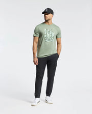 A man in a Psycho Bunny MENS ZEALAND GRAPHIC TEE (B6U604F200) featuring a large Bugs Bunny face graphic, black cap, black pants, and white sneakers stands against a plain white background.
