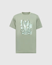 The Psycho Bunny MENS ZEALAND GRAPHIC TEE (B6U604F200) is a light green shirt with a stylized bunny ears and crossed bones graphic on the chest, framed by a subtle rectangular background pattern.
