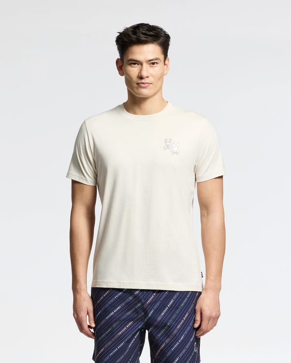A person in a Psycho Bunny MENS ELLIOT FRONT BACK GRAPHIC TEE (B6U605F200), a regular-fit off-white Pima cotton shirt with a small graphic on the left, paired with patterned blue shorts, stands against a light gray background. They have short dark hair and maintain a neutral expression.