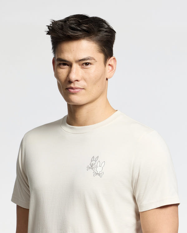 A person with short dark hair is wearing the MENS ELLIOT FRONT BACK GRAPHIC TEE by Psycho Bunny, a regular-fit Pima cotton T-shirt featuring a small outlined cartoon character on a plain white background.