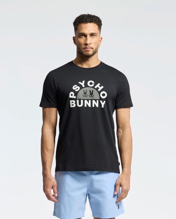A man, wearing the Psycho Bunny Goldie Graphic Tee (B6U606F200) made from Peruvian Pima cotton and featuring the iconic bunny logo, faces forward. He pairs it with light blue shorts against a plain white backdrop.