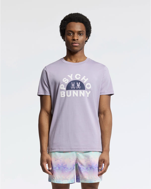 A model displays the MENS GOLDIE GRAPHIC TEE - B6U606F200 by Psycho Bunny, featuring vibrant bunny graphics in luxurious Peruvian Pima cotton, paired with pastel tie-dye shorts on a plain white background.