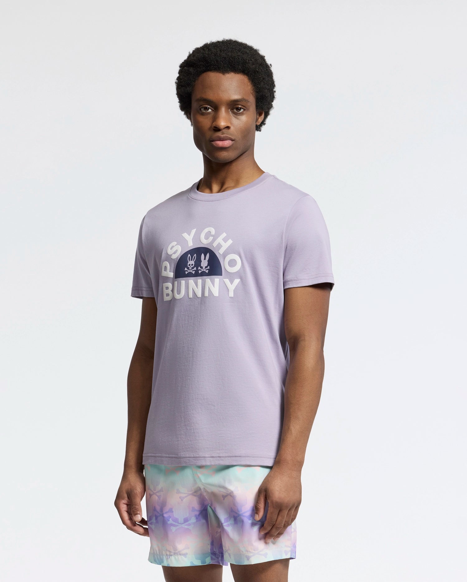 A man wears a Psycho Bunny MENS GOLDIE GRAPHIC TEE - B6U606F200 in light purple, crafted from luxurious Peruvian Pima cotton, featuring a unique bunny graphic. He pairs it with vibrant pastel shorts against a plain white backdrop.
