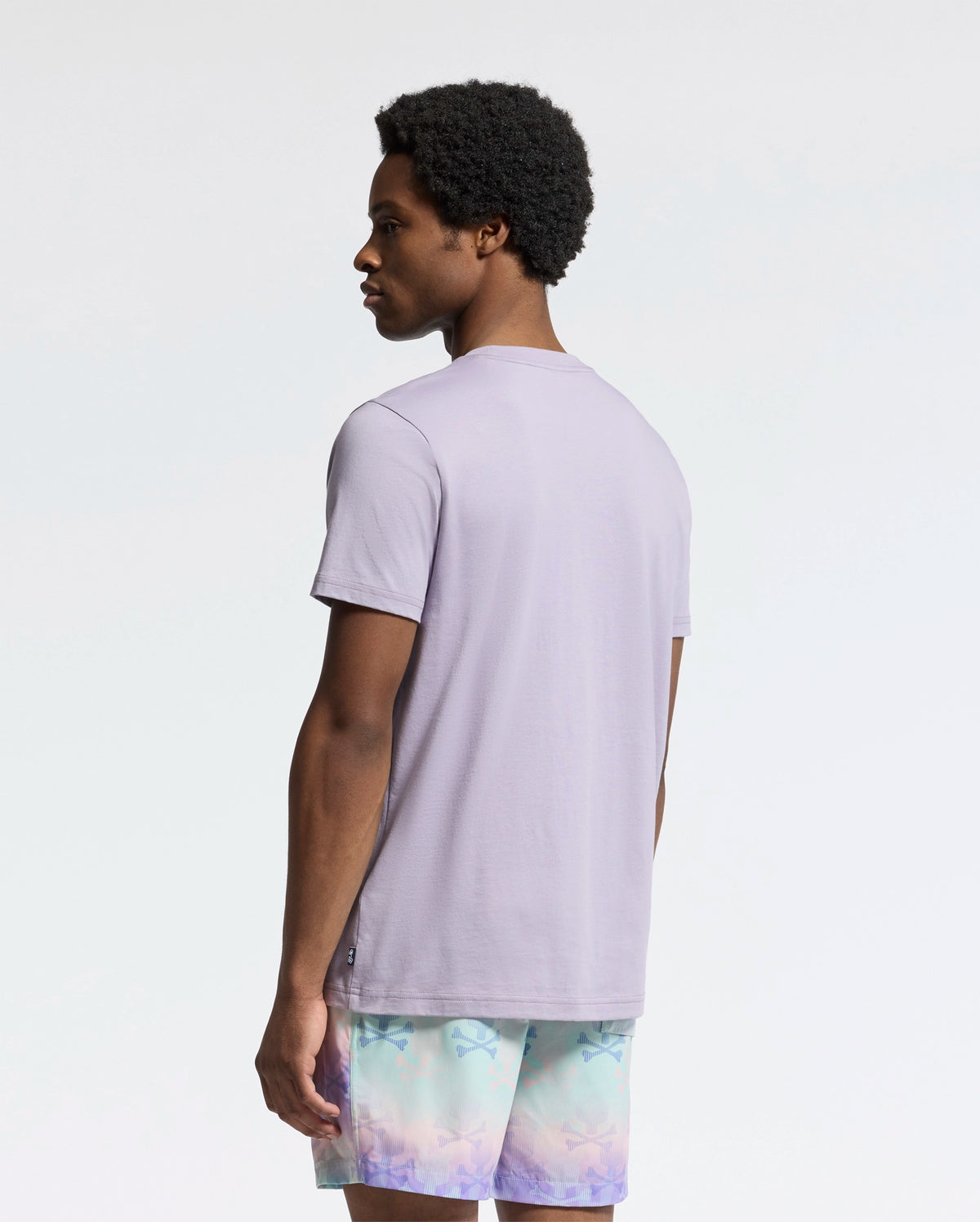 A person with short curly hair stands against a plain background, facing slightly to the side, wearing a Psycho Bunny MENS GOLDIE GRAPHIC TEE in light purple, crafted from Peruvian Pima cotton, paired with pastel gradient pattern shorts.