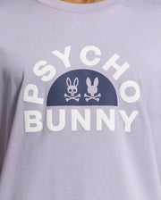 The Psycho Bunny Men's Goldie Graphic Tee (B6U606F200) is crafted from Peruvian Pima cotton and features a light purple color with "Psycho Bunny" in white. It includes a bold graphic of two skull-and-crossbones bunnies—one outlined, one filled—against a dark oval background.