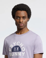 A person with short curly hair wears a light purple MENS GOLDIE GRAPHIC TEE - B6U606F200 crafted from Peruvian Pima cotton by Psycho Bunny, featuring a stylized bunny graphic on the front. They gaze directly at the camera against a plain, light background.