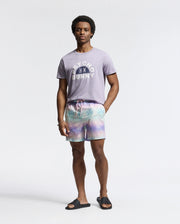 A man in a Psycho Bunny MENS GOLDIE GRAPHIC TEE, crafted from Peruvian Pima cotton and featuring a subtle Bunny graphic, poses against a plain backdrop. He sports pastel tie-dye shorts and black sandals, smiling slightly with one arm relaxed by his side and the other in his pocket.