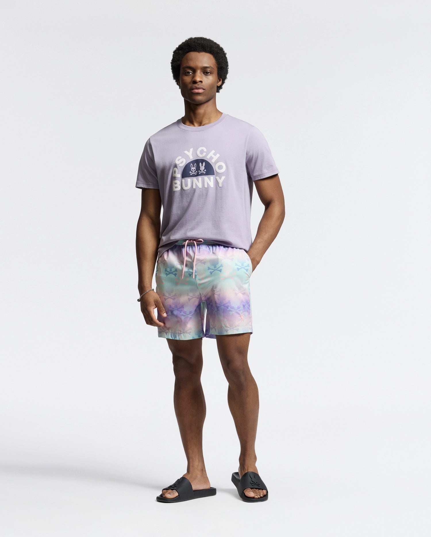 A man in a Psycho Bunny MENS GOLDIE GRAPHIC TEE, crafted from Peruvian Pima cotton and featuring a subtle Bunny graphic, poses against a plain backdrop. He sports pastel tie-dye shorts and black sandals, smiling slightly with one arm relaxed by his side and the other in his pocket.