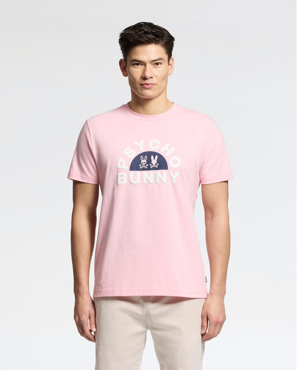 A man poses against a plain backdrop wearing the light pink MENS GOLDIE GRAPHIC TEE - B6U606F200 by Psycho Bunny, adorned with the iconic Bunny Graphic and logo. He complements it with light-colored pants for an effortlessly casual vibe.