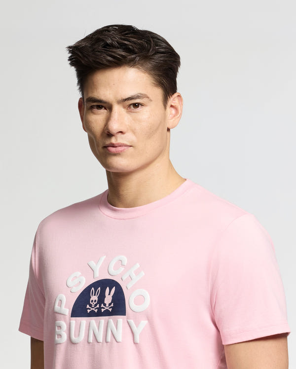 A short dark-haired person models the Goldie Graphic Tee (B6U606F200) by Psycho Bunny in light pink, spotlighting a bold Bunny Graphic with "Psycho Bunny" printed in white on blue. They stand against a light gray backdrop that highlights the Pima Cotton's softness and stylishness.