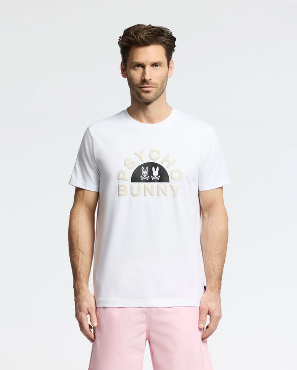 A man confidently stands against a white background in a Psycho Bunny MENS GOLDIE GRAPHIC TEE (B6U606F200), showcasing a bold bunny graphic with crossbones, crafted from soft Pima cotton. He pairs it stylishly with pink shorts.