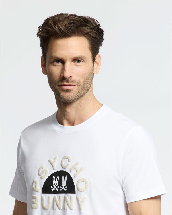 A person with brown hair and a slight beard wears the MENS GOLDIE GRAPHIC TEE (B6U606F200) in white Pima cotton, featuring Psycho Bunny's unique bunny logo. The simple, light-colored background highlights the tee's design.
