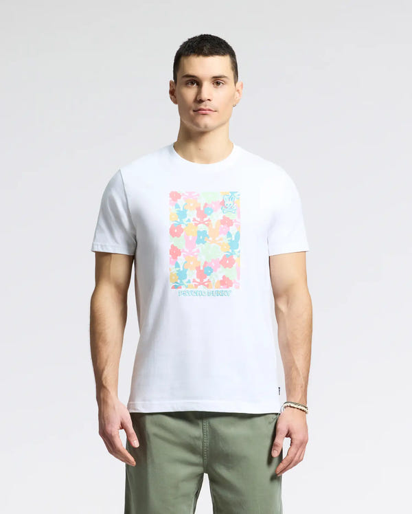 A person exudes casual charm in a Psycho Bunny MENS LEMMON GRAPHIC TEE - B6U655F200. This white tee, with its colorful floral pattern and soft Pima cotton fabric, pairs perfectly with light green pants. They have short hair and stand confidently against a plain background.