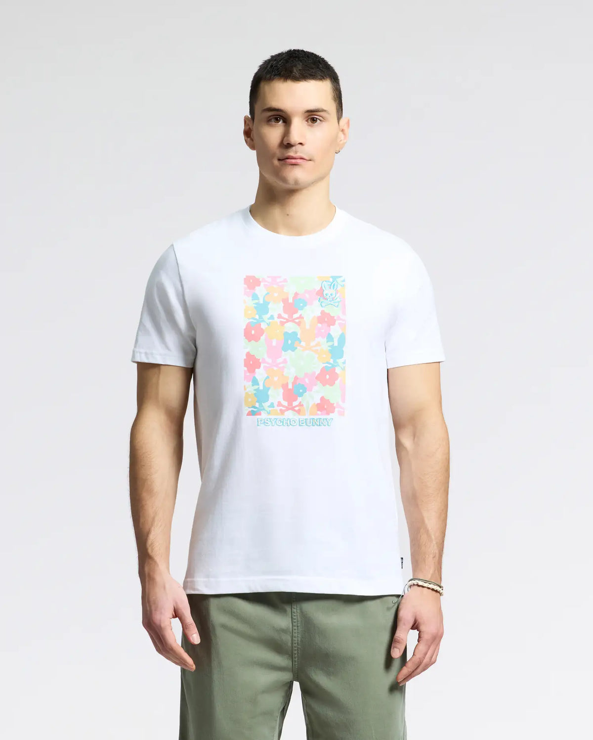 A person exudes casual charm in a Psycho Bunny MENS LEMMON GRAPHIC TEE - B6U655F200. This white tee, with its colorful floral pattern and soft Pima cotton fabric, pairs perfectly with light green pants. They have short hair and stand confidently against a plain background.