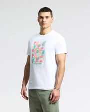 A person stands against a plain background sporting a Psycho Bunny MENS LEMMON GRAPHIC TEE - B6U655F200, a white Pima cotton shirt with a colorful abstract design. They have short dark hair and complement the look with light green pants, creating a comfortable style.