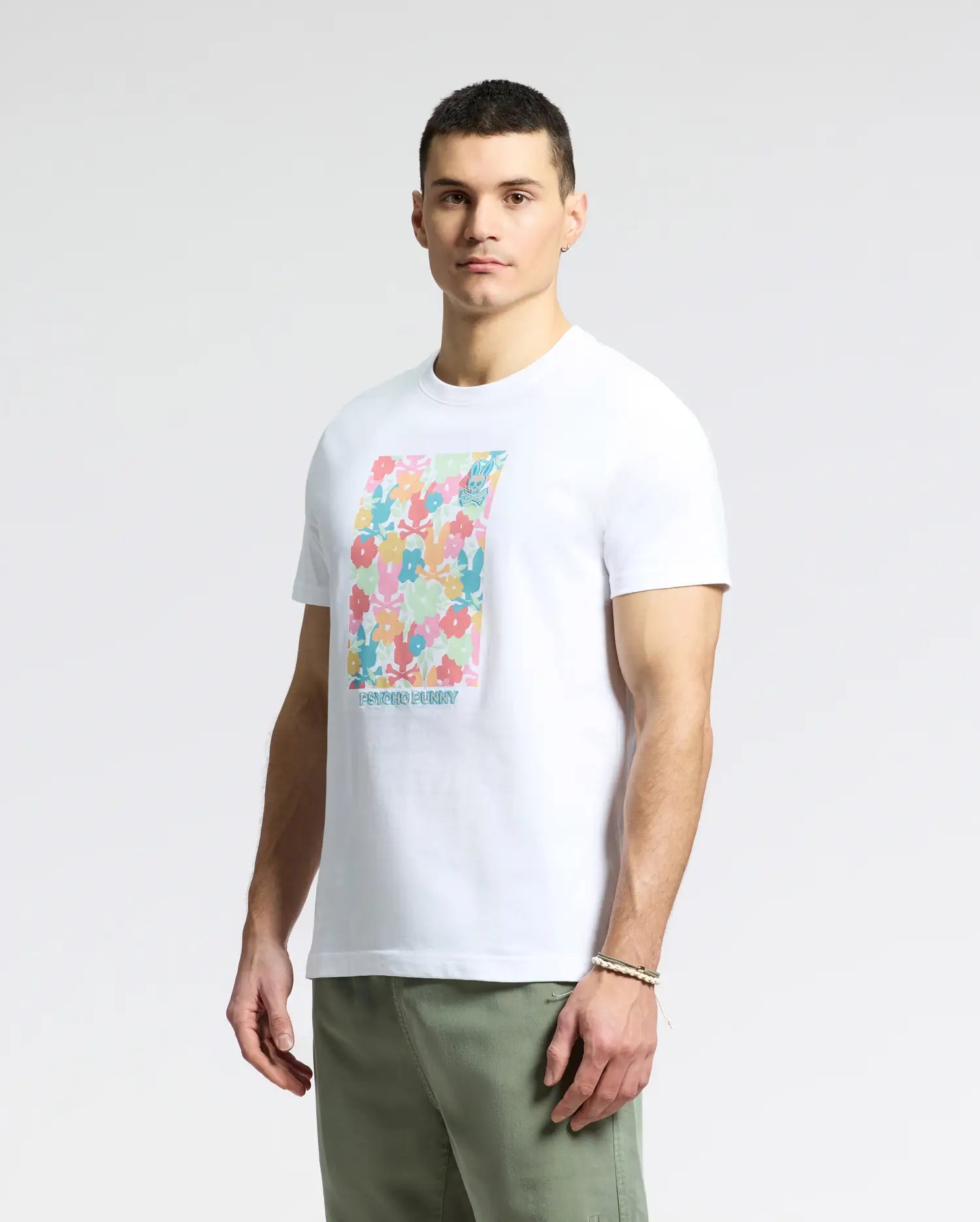 A person stands against a plain background sporting a Psycho Bunny MENS LEMMON GRAPHIC TEE - B6U655F200, a white Pima cotton shirt with a colorful abstract design. They have short dark hair and complement the look with light green pants, creating a comfortable style.