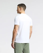 A person with short dark hair stands sideways, looking right. They're wearing the MENS LEMMON GRAPHIC TEE by Psycho Bunny and light green pants. The comfortable tee in Pima cotton beautifully complements the neutral background.