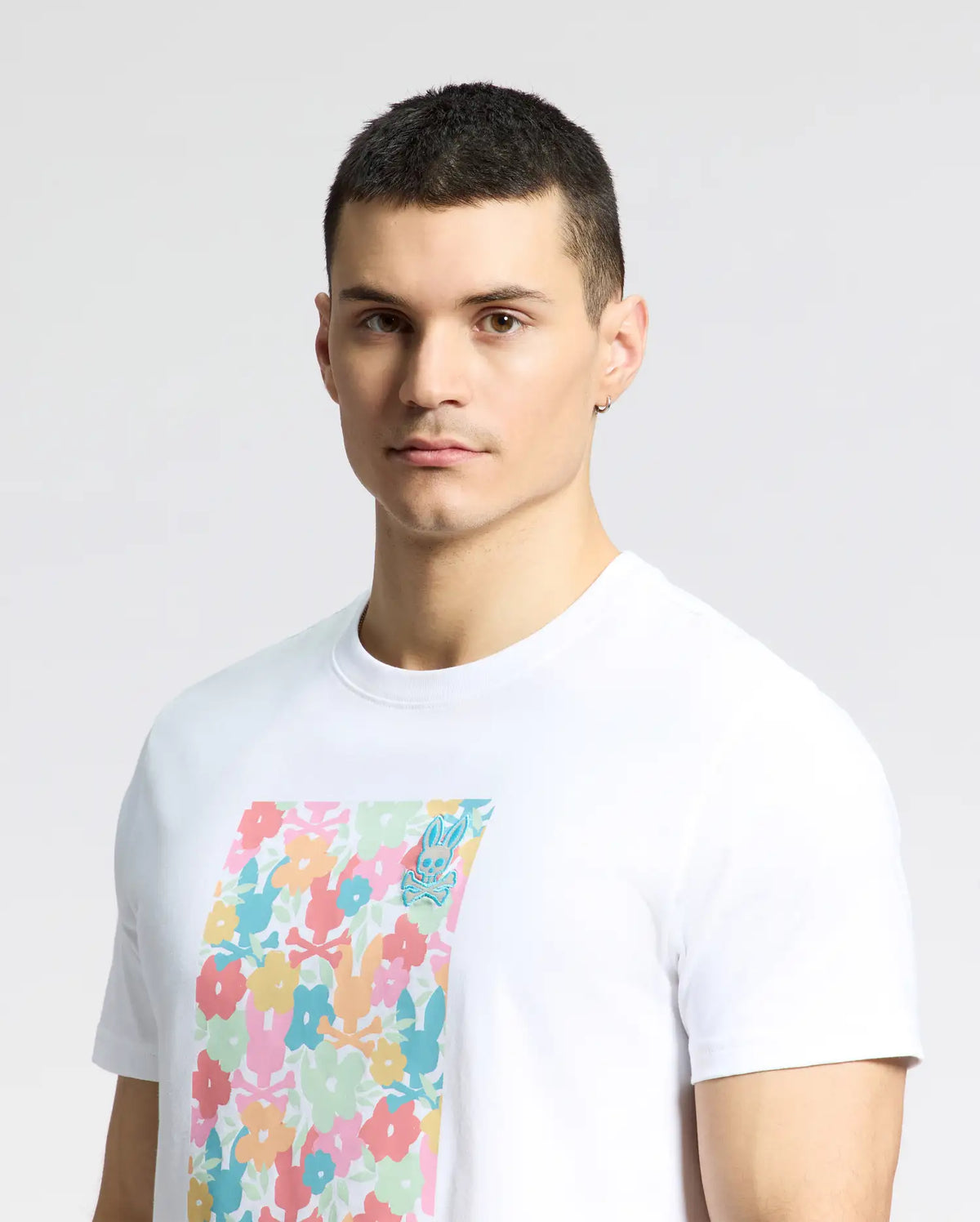 A person with short dark hair wears the Psycho Bunny MENS LEMMON GRAPHIC TEE - B6U655F200, a white Pima cotton t-shirt with a vibrant floral design on the front. The light gray background complements their comfortable style.