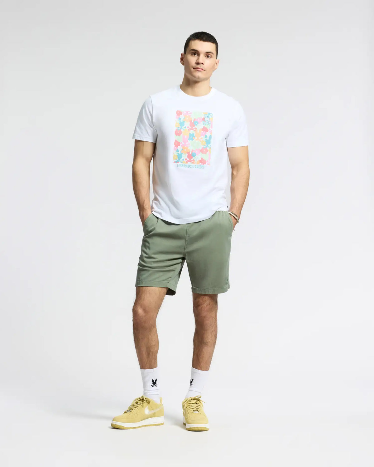 A person stands against a gray background wearing a Psycho Bunny MENS LEMMON GRAPHIC TEE in white Pima cotton with an abstract design, olive green shorts, white socks, and yellow sneakers. Their hands are in their pockets.
