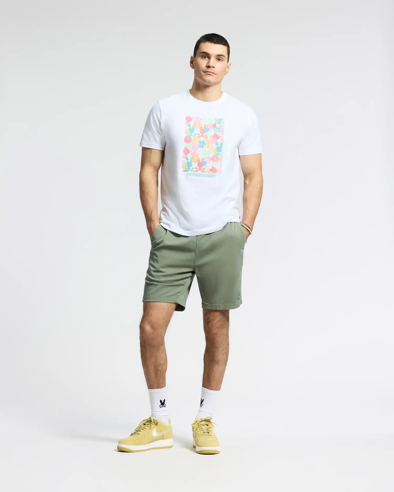 A person stands against a gray background wearing a Psycho Bunny MENS LEMMON GRAPHIC TEE in white Pima cotton with an abstract design, olive green shorts, white socks, and yellow sneakers. Their hands are in their pockets.
