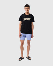 A young man smiling, dressed in a MENS MELVIN GRAPHIC TEE - B6U663C200 from Psycho Bunny, made of Pima cotton with "Psycho Bunny" in HD-silicone print and a bunny logo on it, paired with light blue shorts and black slide sandals, stands against a plain gray background.