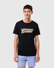 A person with short black hair smiles while wearing the MENS MELVIN GRAPHIC TEE - B6U663C200 by Psycho Bunny, a regular fit black T-shirt made of Pima cotton featuring the brand’s beige logo with an HD-silicone print bunny. The person is also wearing light purple shorts, set against a plain white background.