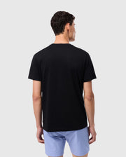 A person with short dark hair stands facing away from the camera, wearing a Psycho Bunny MENS MELVIN GRAPHIC TEE - B6U663C200 made of Pima cotton and light blue shorts. The background is plain and light-colored.