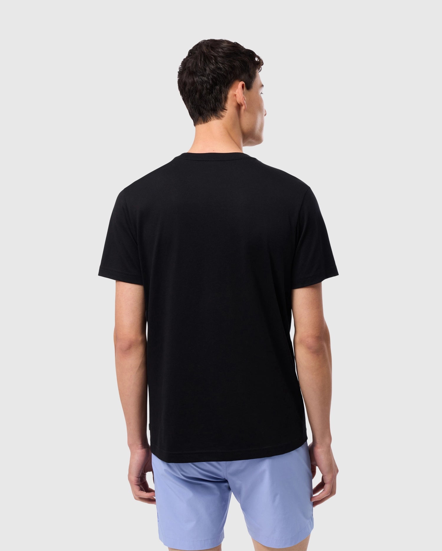 A person with short dark hair stands facing away from the camera, wearing a Psycho Bunny MENS MELVIN GRAPHIC TEE - B6U663C200 made of Pima cotton and light blue shorts. The background is plain and light-colored.