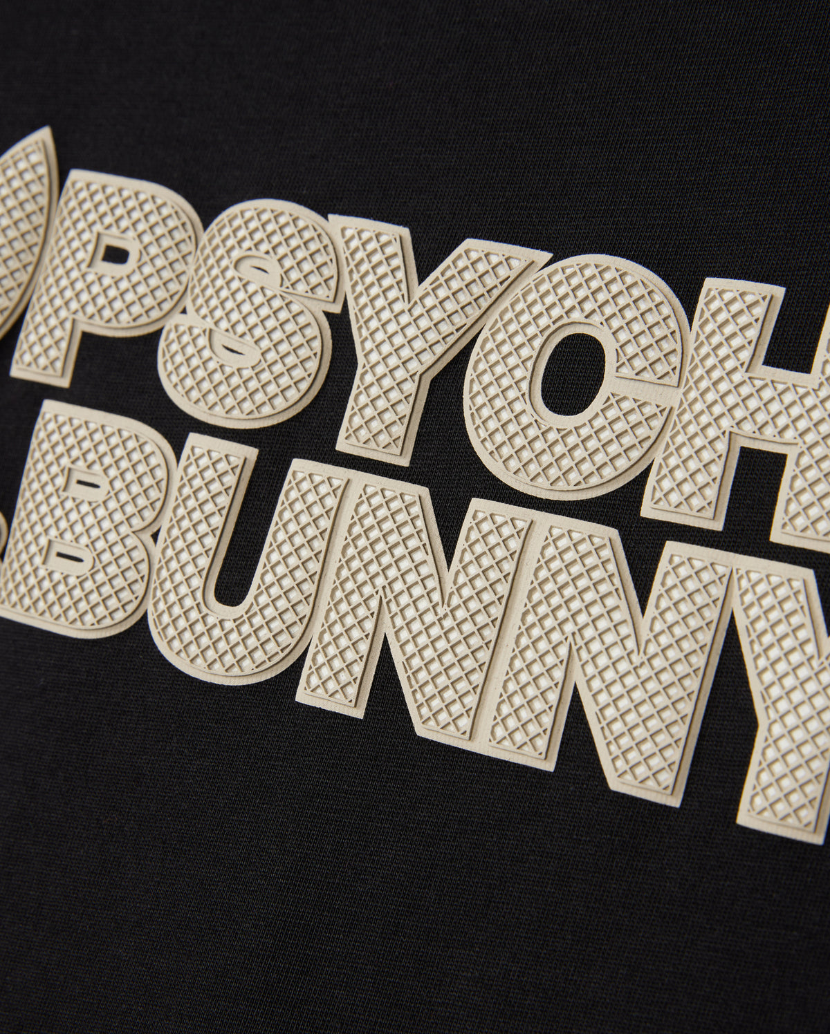 Close-up of the MENS MELVIN GRAPHIC TEE - B6U663C200 from Psycho Bunny, showcasing black fabric with "PSYCHO BUNNY" prominently displayed in large, textured, cream-colored letters. The distinctive HD-silicone print adds a three-dimensional effect to the text. Made from Pima cotton, this tee ensures both comfort and durability.