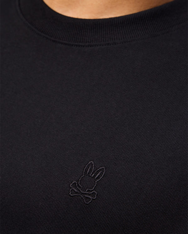 Close-up of a person wearing the Psycho Bunny MENS DUKE OVERSIZED TEE - B6U742D200 featuring an embroidered bunny skull and crossbones logo. Crafted from Pima cotton, this black shirt boasts a ribbed neckline and smooth fabric texture. The image focuses on the torso area, highlighting both the relaxed fit and the intricate logo detail.