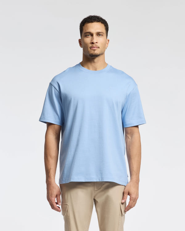 A man with short hair and a neutral expression stands against a plain background wearing a Psycho Bunny MENS DUKE OVERSIZED TEE in light blue, crafted from premium Pima cotton, paired with beige pants.