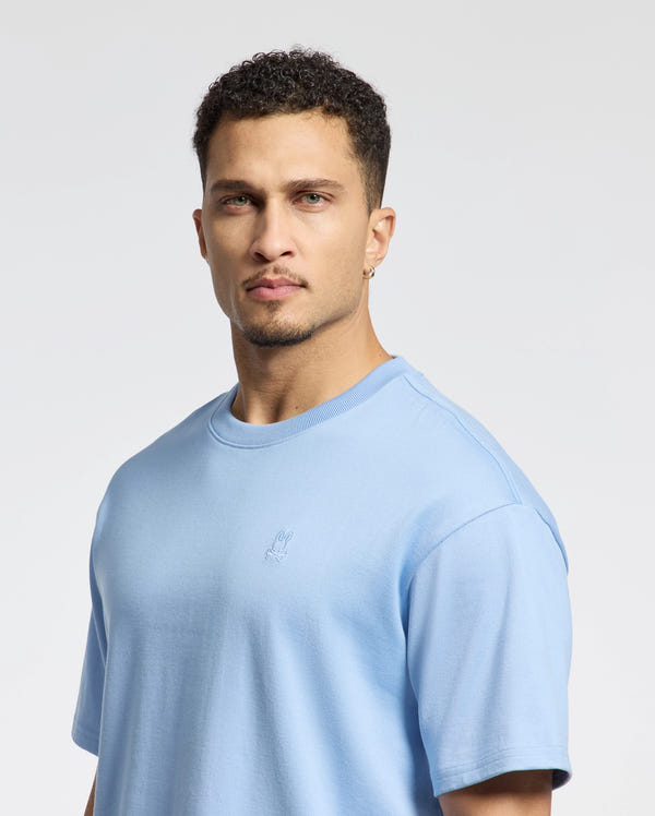 A man with short curly hair and an earring wears a Psycho Bunny MENS DUKE OVERSIZED TEE - B6U742D200, a relaxed fit in light blue Pima cotton. He gazes directly at the camera against a plain, light-colored background.