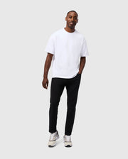 A man is standing against a plain, light grey background. He is wearing a loose-fitting white MENS DUKE OVERSIZED TEE - B6U742D200 by Psycho Bunny, black pants, and white sneakers with black accents. The man has a relaxed posture, with one hand in his pocket and is smiling slightly.