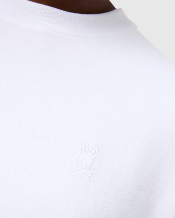 Close-up of the MENS DUKE OVERSIZED TEE - B6U742D200 from Psycho Bunny, a white heavyweight Pima cotton T-shirt featuring an embroidered bunny design with crossed bones underneath. The background is neutral grey, showcasing part of the person’s shoulder and neckline to emphasize the relaxed fit and solid pattern of the garment.