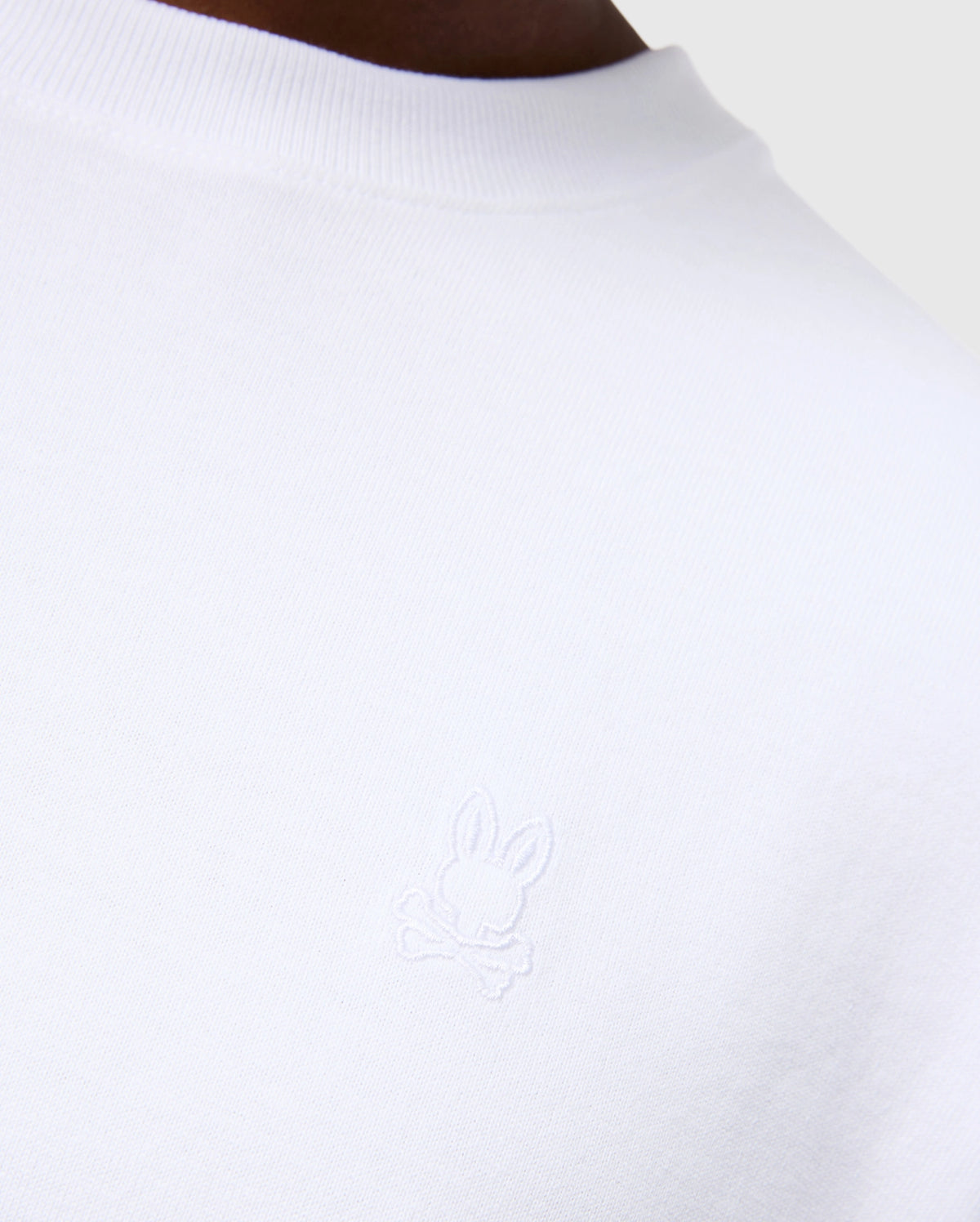Close-up of the MENS DUKE OVERSIZED TEE - B6U742D200 from Psycho Bunny, a white heavyweight Pima cotton T-shirt featuring an embroidered bunny design with crossed bones underneath. The background is neutral grey, showcasing part of the person’s shoulder and neckline to emphasize the relaxed fit and solid pattern of the garment.