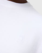 Close-up of the MENS DUKE OVERSIZED TEE - B6U742D200 from Psycho Bunny, a white heavyweight Pima cotton T-shirt featuring an embroidered bunny design with crossed bones underneath. The background is neutral grey, showcasing part of the person’s shoulder and neckline to emphasize the relaxed fit and solid pattern of the garment.