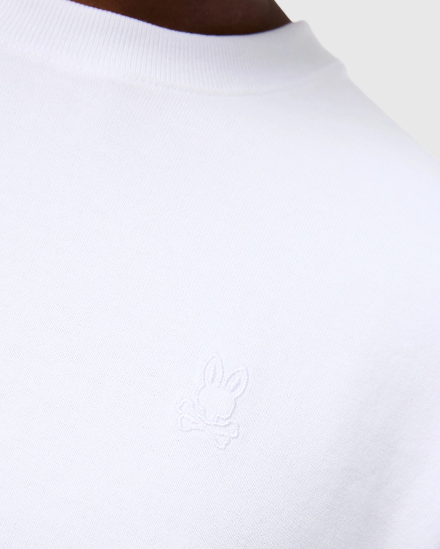 Close-up of the MENS DUKE OVERSIZED TEE - B6U742D200 from Psycho Bunny, a white heavyweight Pima cotton T-shirt featuring an embroidered bunny design with crossed bones underneath. The background is neutral grey, showcasing part of the person’s shoulder and neckline to emphasize the relaxed fit and solid pattern of the garment.