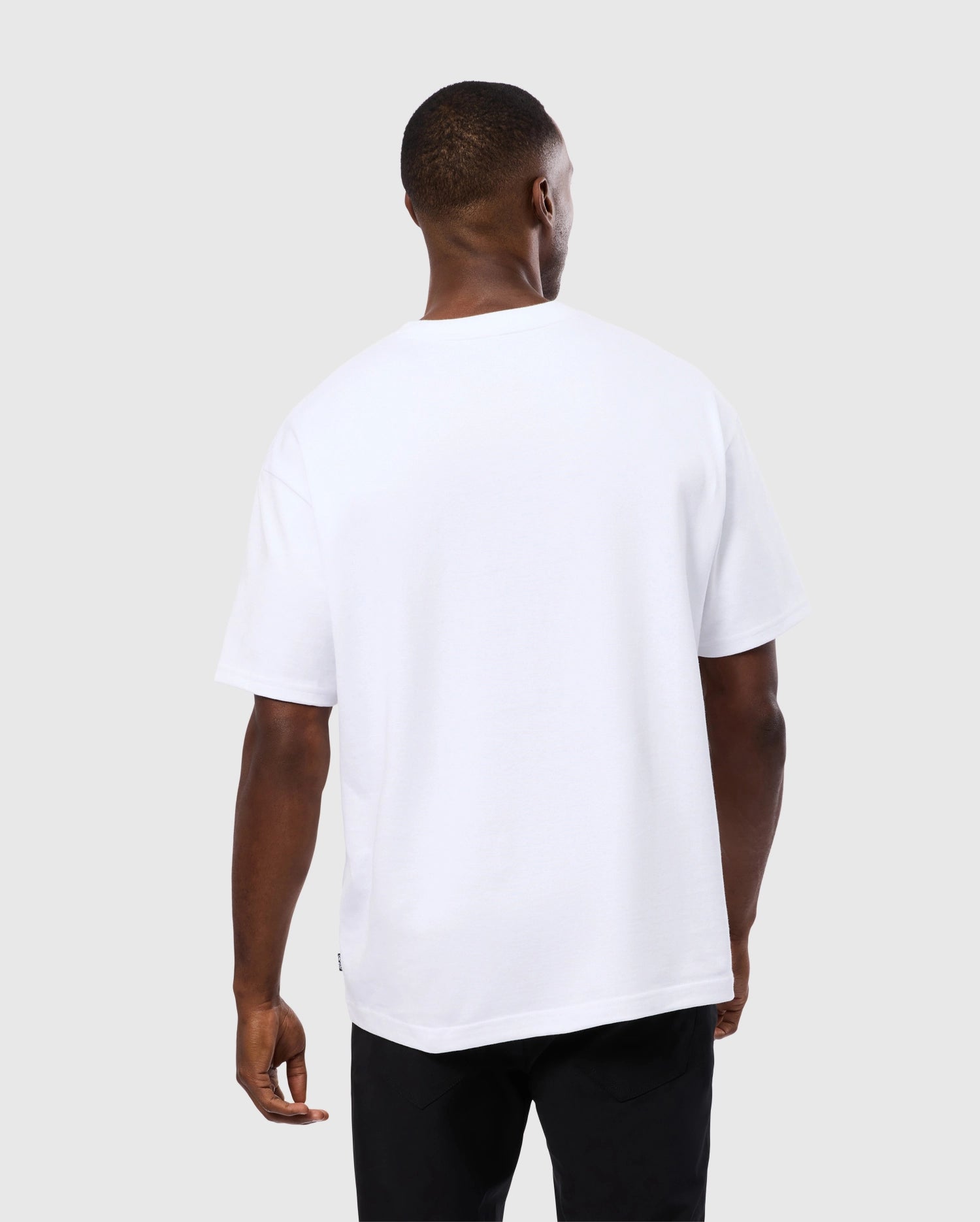 A person with short black hair is standing with their back to the camera, wearing a simple, white MENS DUKE OVERSIZED TEE - B6U742D200 from Psycho Bunny. The short-sleeved t-shirt made of heavyweight Pima cotton and paired with black pants features a relaxed fit and solid pattern, enhancing its casual style. The background is plain and light gray.