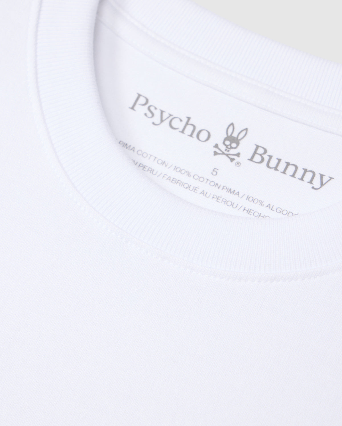 Close-up of a white Psycho Bunny t-shirt label. The label displays the brand name "Psycho Bunny," a rabbit logo with crossbones, and indicates the shirt is made from 100% heavyweight Pima cotton and manufactured in Peru. The background of the image is the solid pattern fabric of this relaxed fit MENS DUKE OVERSIZED TEE - B6U742D200.