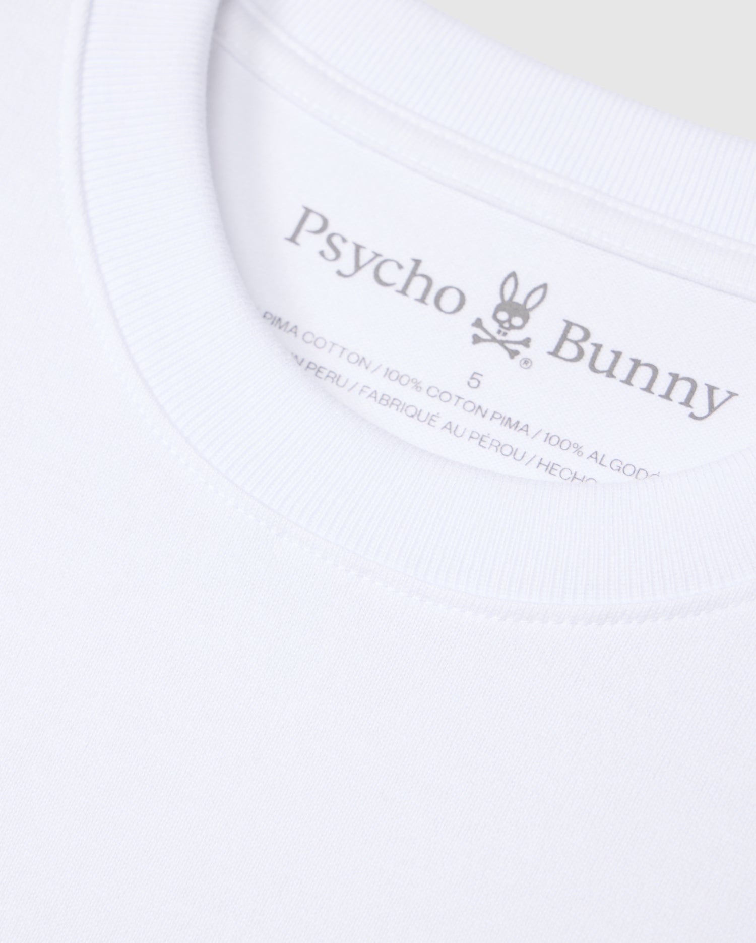 Close-up of a white Psycho Bunny t-shirt label. The label displays the brand name "Psycho Bunny," a rabbit logo with crossbones, and indicates the shirt is made from 100% heavyweight Pima cotton and manufactured in Peru. The background of the image is the solid pattern fabric of this relaxed fit MENS DUKE OVERSIZED TEE - B6U742D200.
