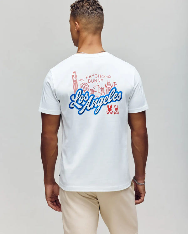 A person showcases the MENS LOS ANGELES CITY TEE (B6U861A2PC) by Psycho Bunny, featuring red line art of Los Angeles landmarks on white Pima cotton. The casual Dodgers style is complete with beige pants, complementing their short curly hair.