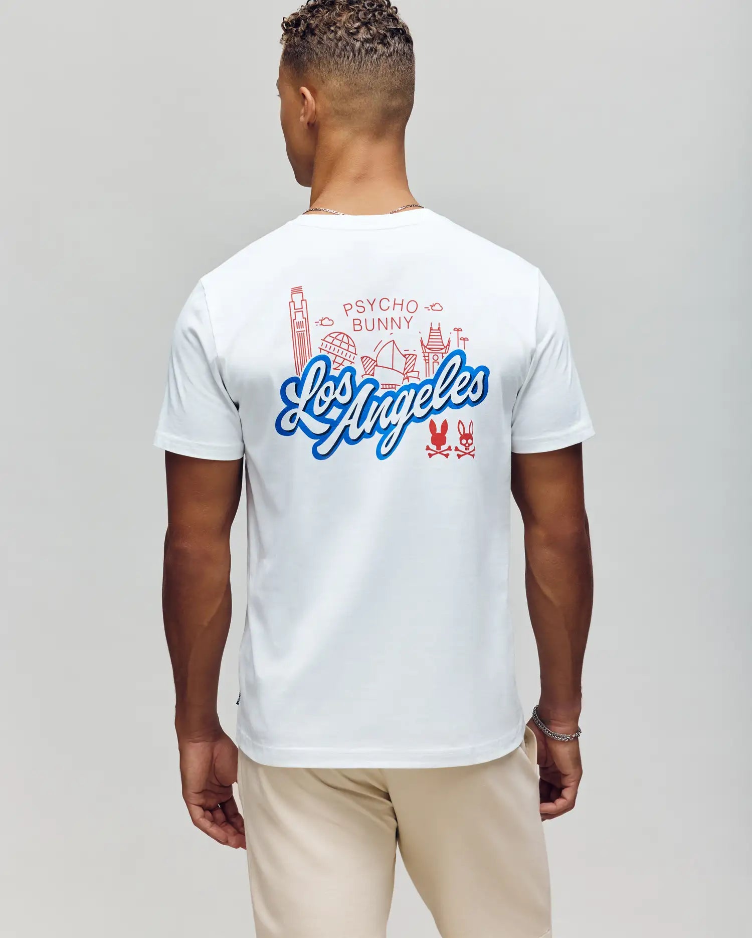 A person showcases the MENS LOS ANGELES CITY TEE (B6U861A2PC) by Psycho Bunny, featuring red line art of Los Angeles landmarks on white Pima cotton. The casual Dodgers style is complete with beige pants, complementing their short curly hair.