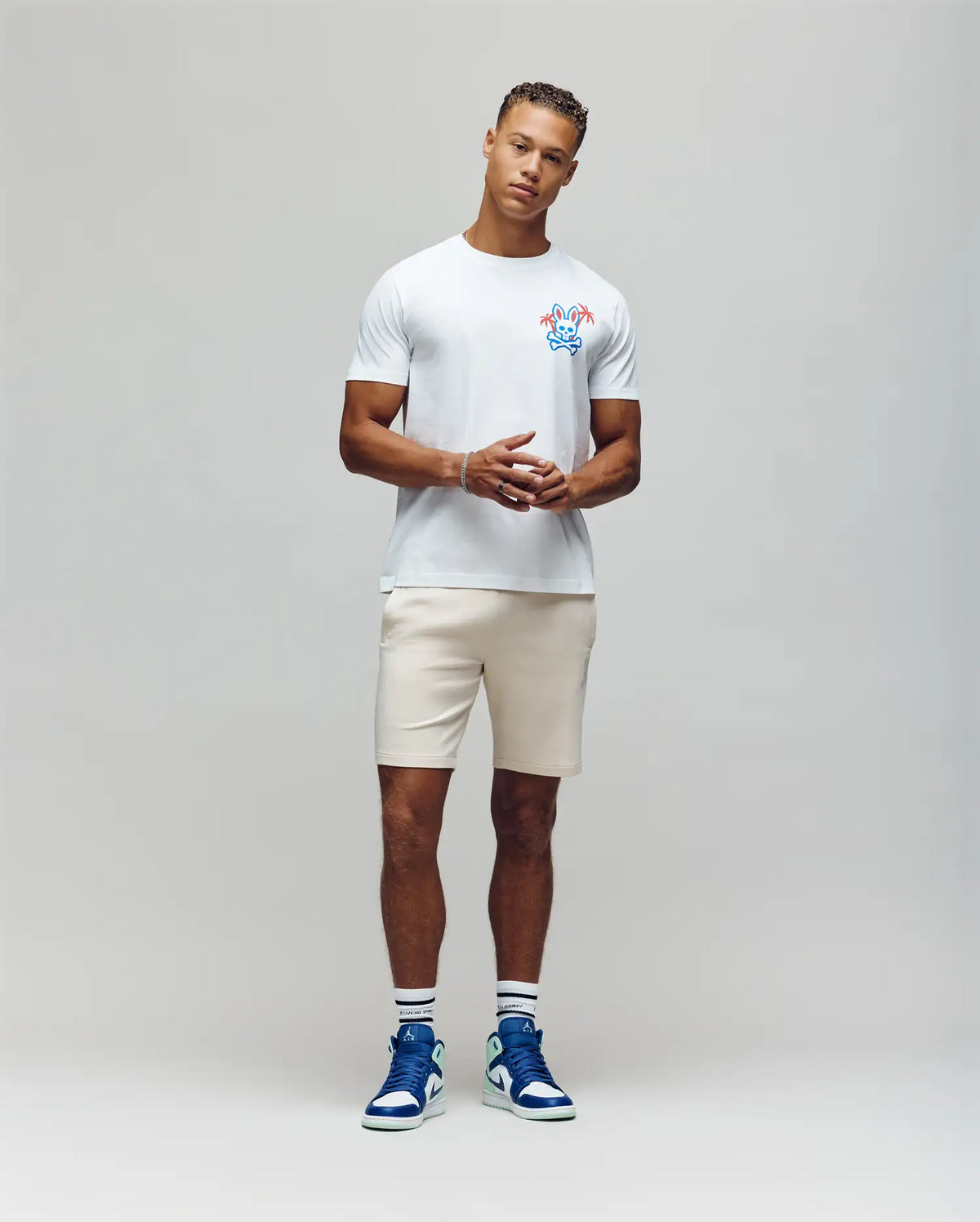 A man poses confidently with clasped hands against a gray background, wearing the Psycho Bunny MENS LOS ANGELES CITY TEE - B6U861A2PC, made of soft Pima cotton and featuring a colorful chest graphic. He's paired it with beige shorts, white socks, and blue-and-white sneakers. He has short curly hair.