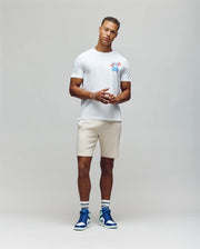 A man poses confidently with clasped hands against a gray background, wearing the Psycho Bunny MENS LOS ANGELES CITY TEE - B6U861A2PC, made of soft Pima cotton and featuring a colorful chest graphic. He's paired it with beige shorts, white socks, and blue-and-white sneakers. He has short curly hair.