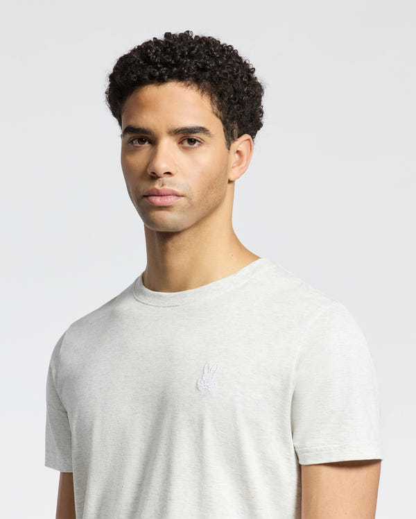 A man with short curly hair wears the light gray Men's James Crew Neck Tee by Psycho Bunny, featuring a small, intricately embroidered bunny logo on the chest, against a plain light background.