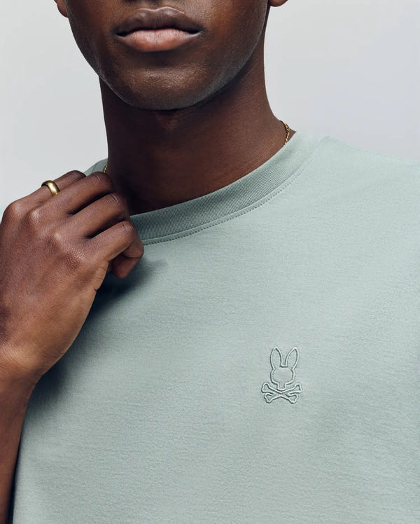 A close-up of a person wearing the Psycho Bunny MENS JAMES PREMIUM CREW NECK TEE showcases the embroidered bunny and crossbones logo, while their gold-ringed hand near the neckline highlights its premium craftsmanship.