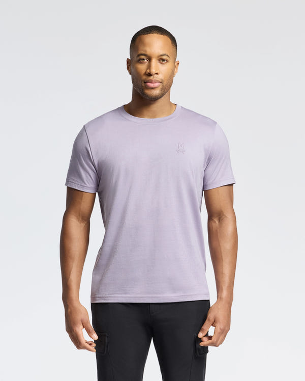 A man wears a Psycho Bunny MENS JAMES CREW NECK TEE - B6U952D200 in light purple with embroidery detail, paired with black pants, set against a plain white background.