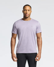 A man wears a Psycho Bunny MENS JAMES CREW NECK TEE - B6U952D200 in light purple with embroidery detail, paired with black pants, set against a plain white background.