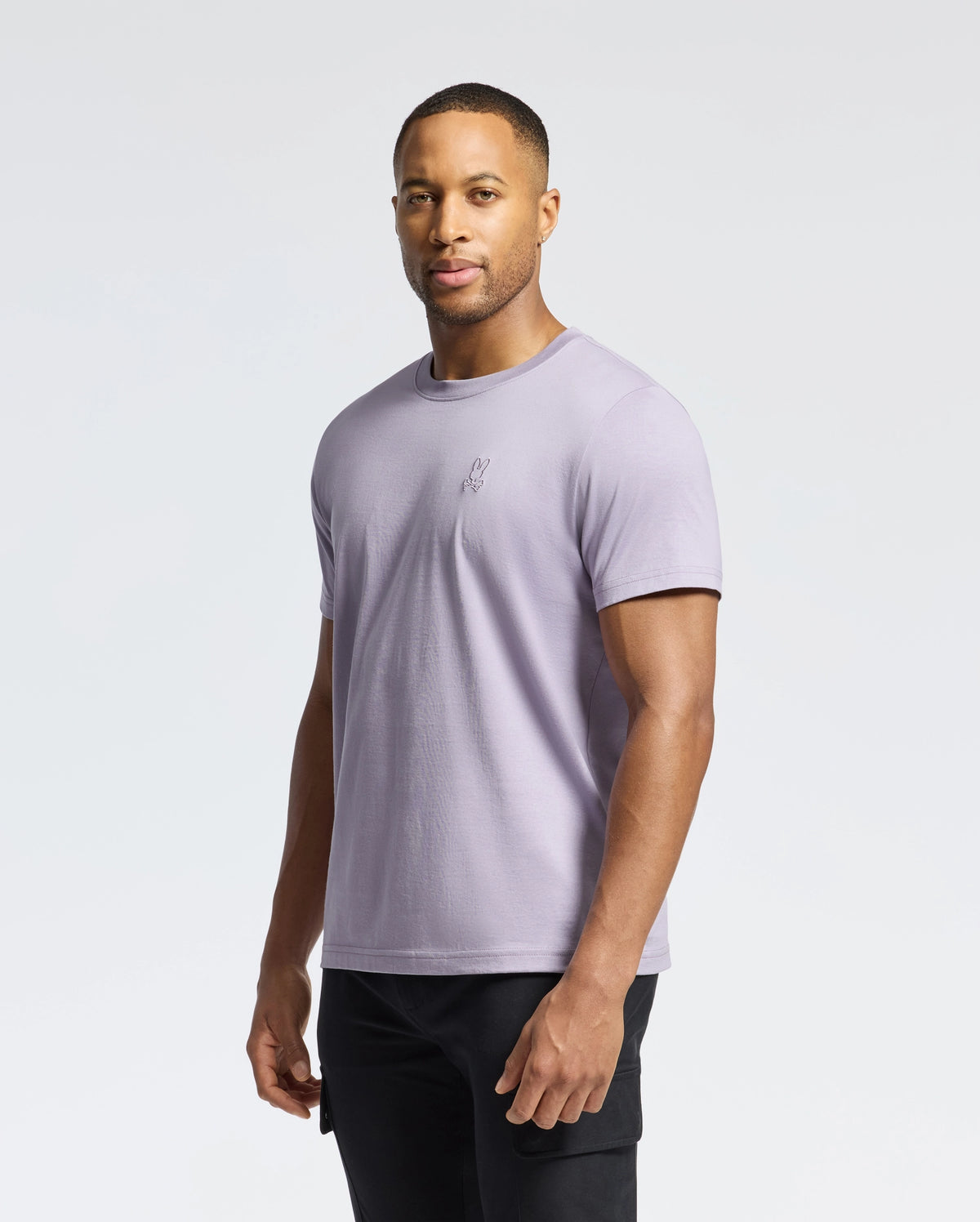 A person in a Psycho Bunny MENS JAMES CREW NECK TEE - B6U952D200, light purple with delicate embroidery, paired with black pants, stands against a plain white background, facing forward neutrally.