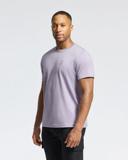 A person in a Psycho Bunny MENS JAMES CREW NECK TEE - B6U952D200, light purple with delicate embroidery, paired with black pants, stands against a plain white background, facing forward neutrally.
